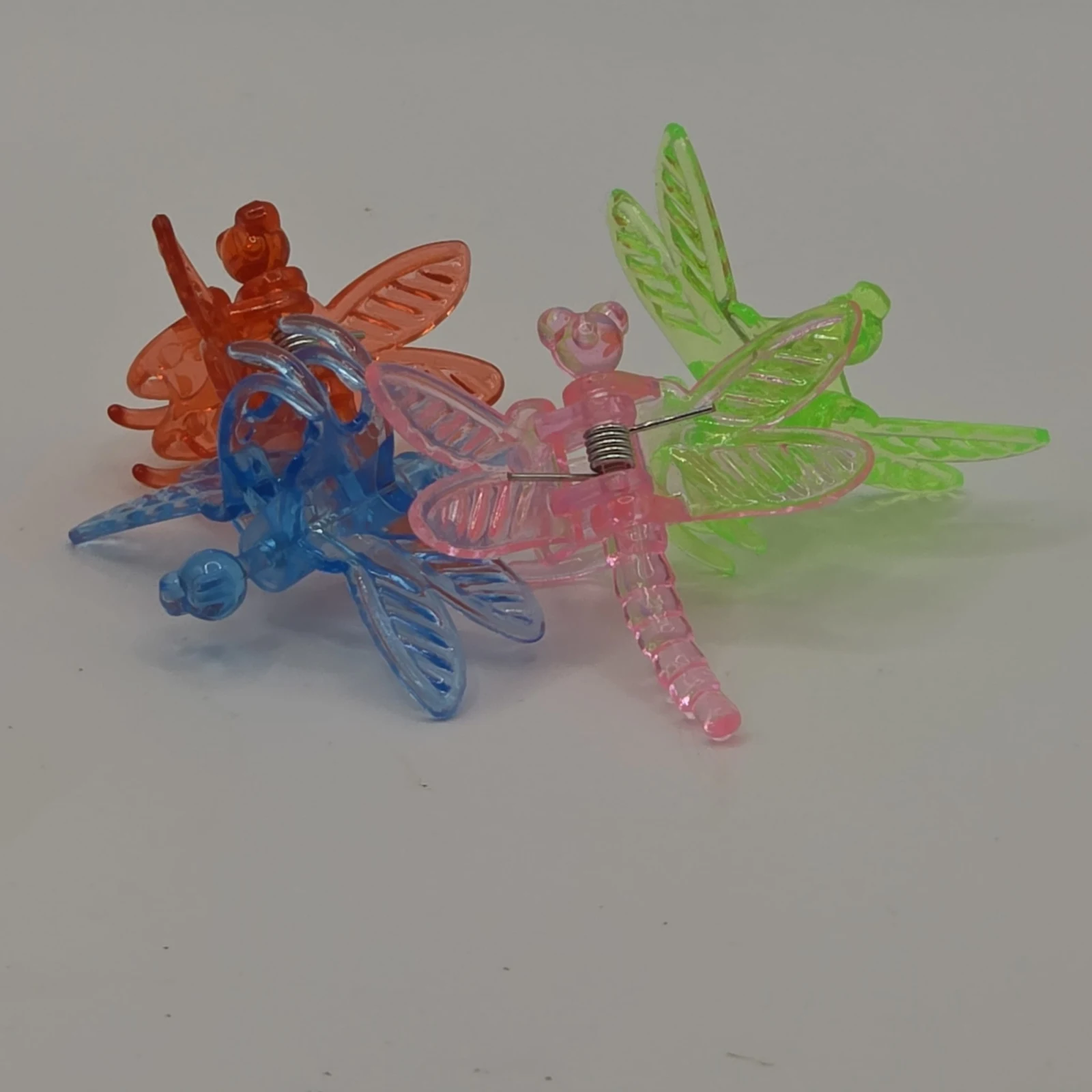 Three translucent dragonfly clips in red, blue, and pink with green wings on white surface.