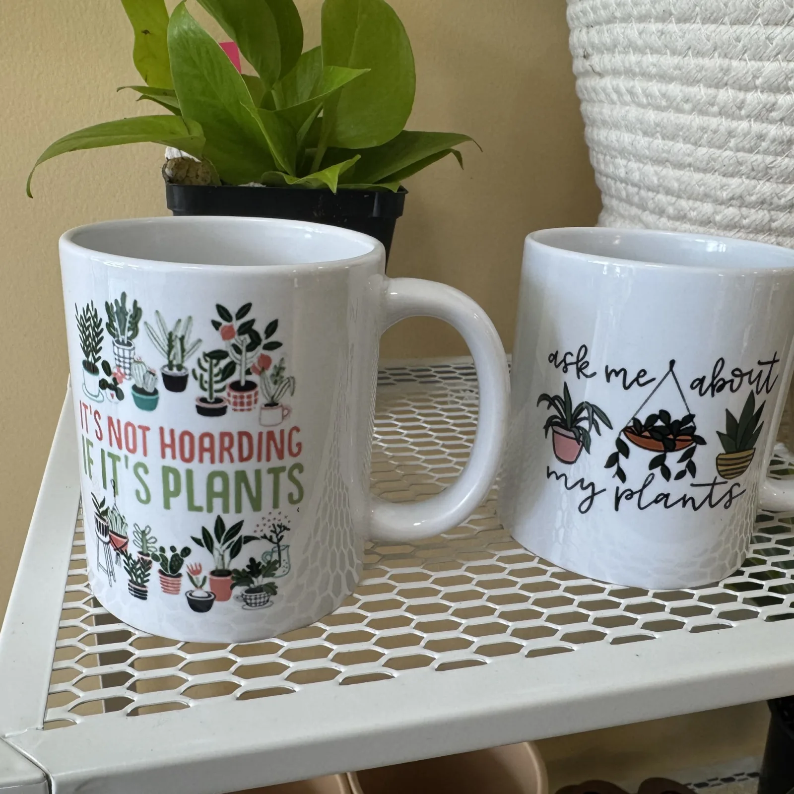 Two white mugs with plant-themed designs displayed on patterned surface with potted plant background.