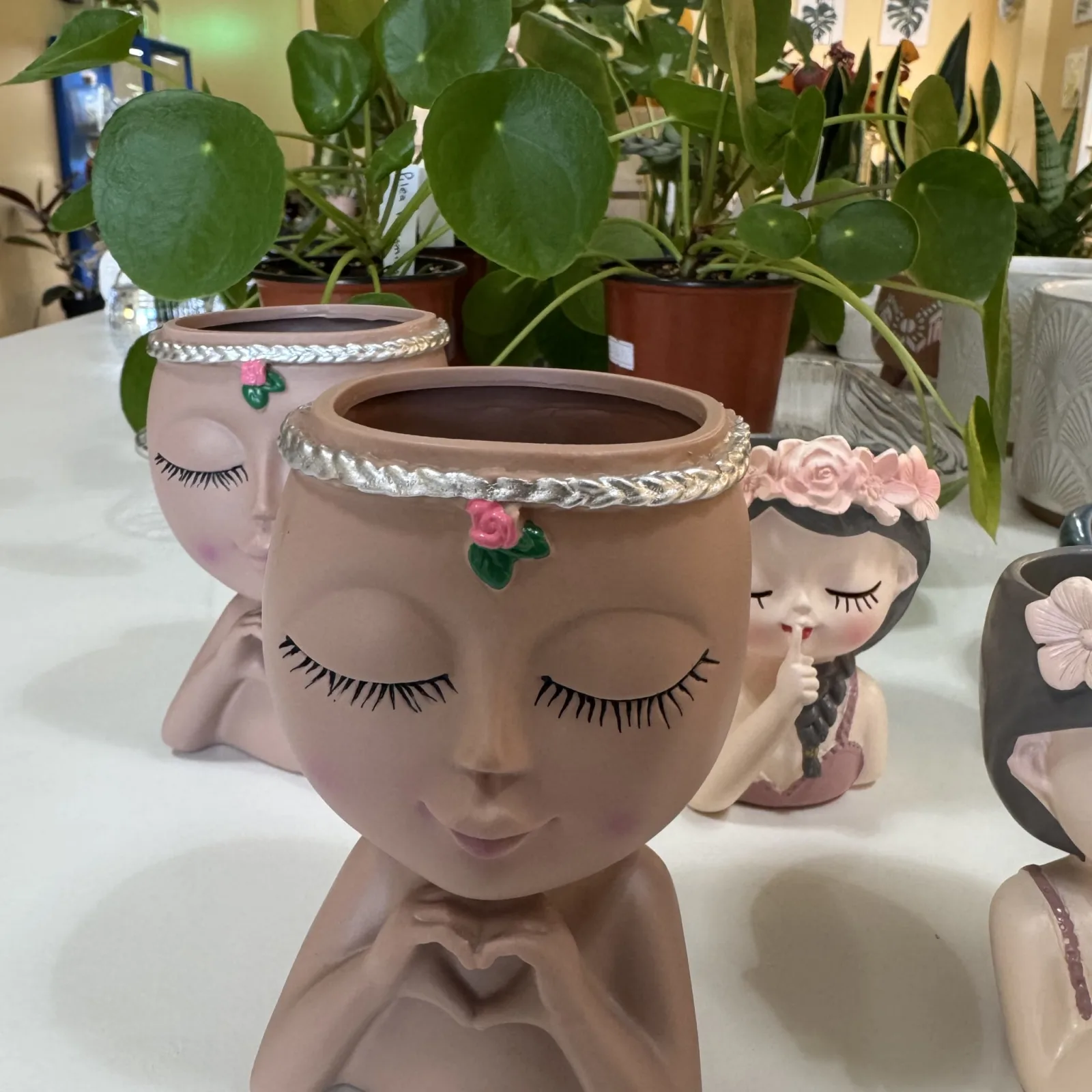Face Plant Pots with closed eyes, pink decorative headband, holding green leafy plants indoors.