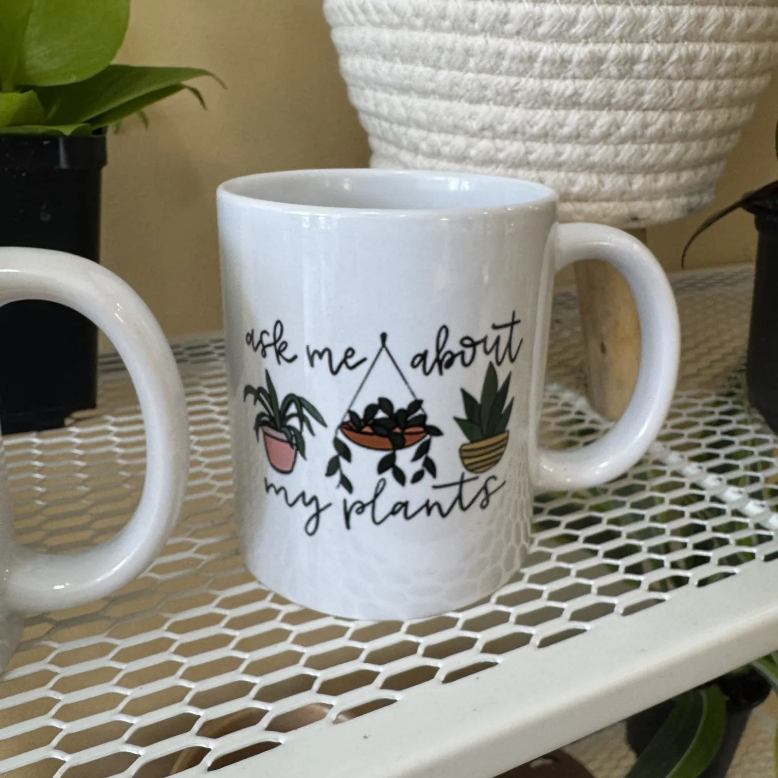 Two white mugs with plant-themed designs displayed on patterned surface with potted plant background.